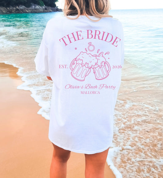 Weißes Bachelorette JGA T-Shirt von Cincerely Yours mit rosa 'The Bride' Aufdruck, clinkenden Bierkrügen und Mallorca Schriftzug, Rückansicht am Strand.