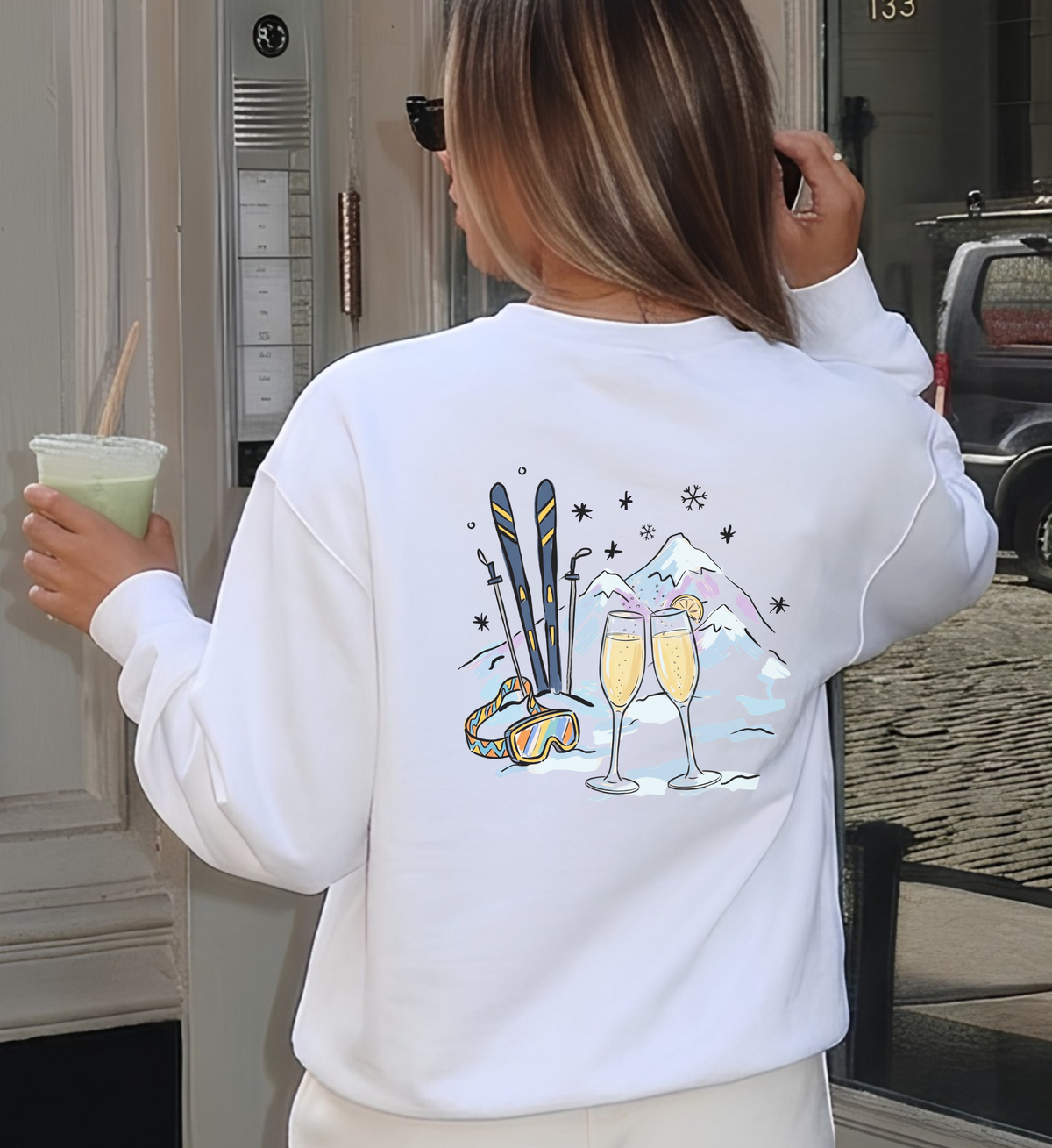 Weißes Après-Ski-Sweatshirt von hinten getragen, mit zwei Mimosa-Gläsern, Skiern, Skibrille und Bergen als Rückenmotiv.
