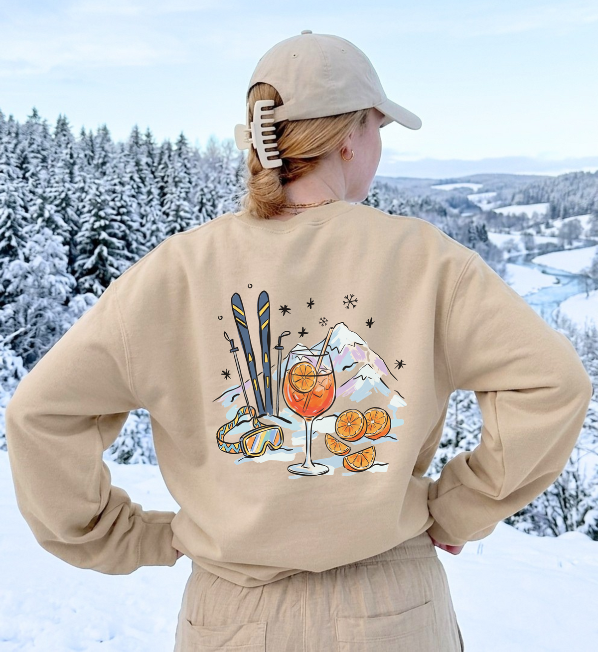 Sandfarbenes Après-Ski-Sweatshirt von hinten mit Spritz-, Orangen- und Skimotiv vor verschneiter Landschaft.
