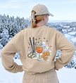 Sandfarbenes Après-Ski-Sweatshirt von hinten mit Spritz-, Orangen- und Skimotiv vor verschneiter Landschaft.
