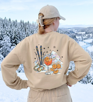 Sandfarbenes Après-Ski-Sweatshirt von hinten mit Spritz-, Orangen- und Skimotiv vor verschneiter Landschaft.
