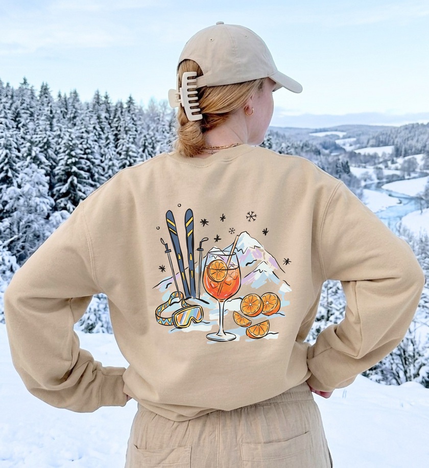 Sandfarbenes Après-Ski-Sweatshirt von hinten mit Spritz-, Orangen- und Skimotiv vor verschneiter Landschaft.
