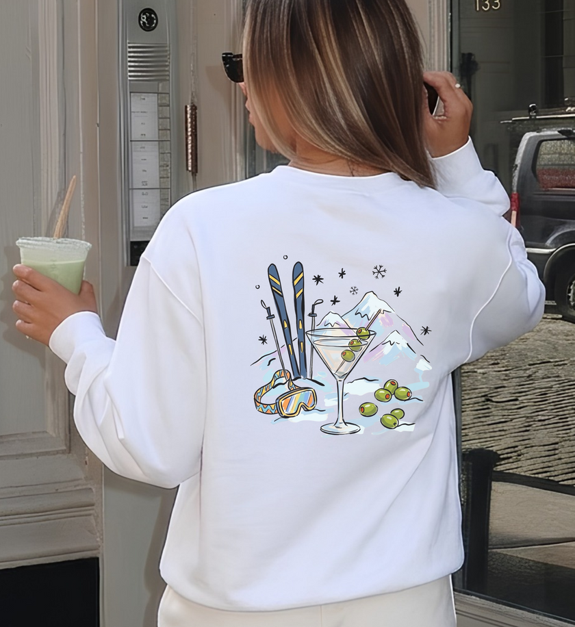 Weißes Après-Ski-Sweatshirt von hinten getragen, mit Martini, Oliven, Skiern, Skibrille und Bergen als Rückenmotiv.


