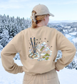 Sandfarbenes Après-Ski-Sweatshirt in Rückansicht mit Martini-, Ski- und Bergmotiv vor verschneiter Landschaft.
