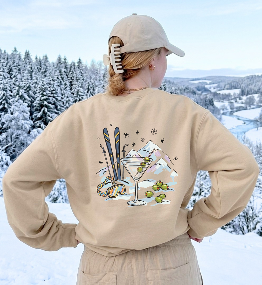 Sandfarbenes Après-Ski-Sweatshirt in Rückansicht mit Martini-, Ski- und Bergmotiv vor verschneiter Landschaft.
