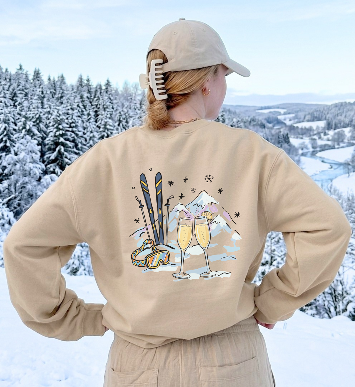 Sandfarbenes Après-Ski-Sweatshirt von hinten getragen, mit zwei Mimosa-Gläsern, Skiern und Bergen vor Schneelandschaft.

