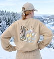 Sandfarbenes Après-Ski-Sweatshirt von hinten getragen, mit zwei Mimosa-Gläsern, Skiern und Bergen vor Schneelandschaft.

