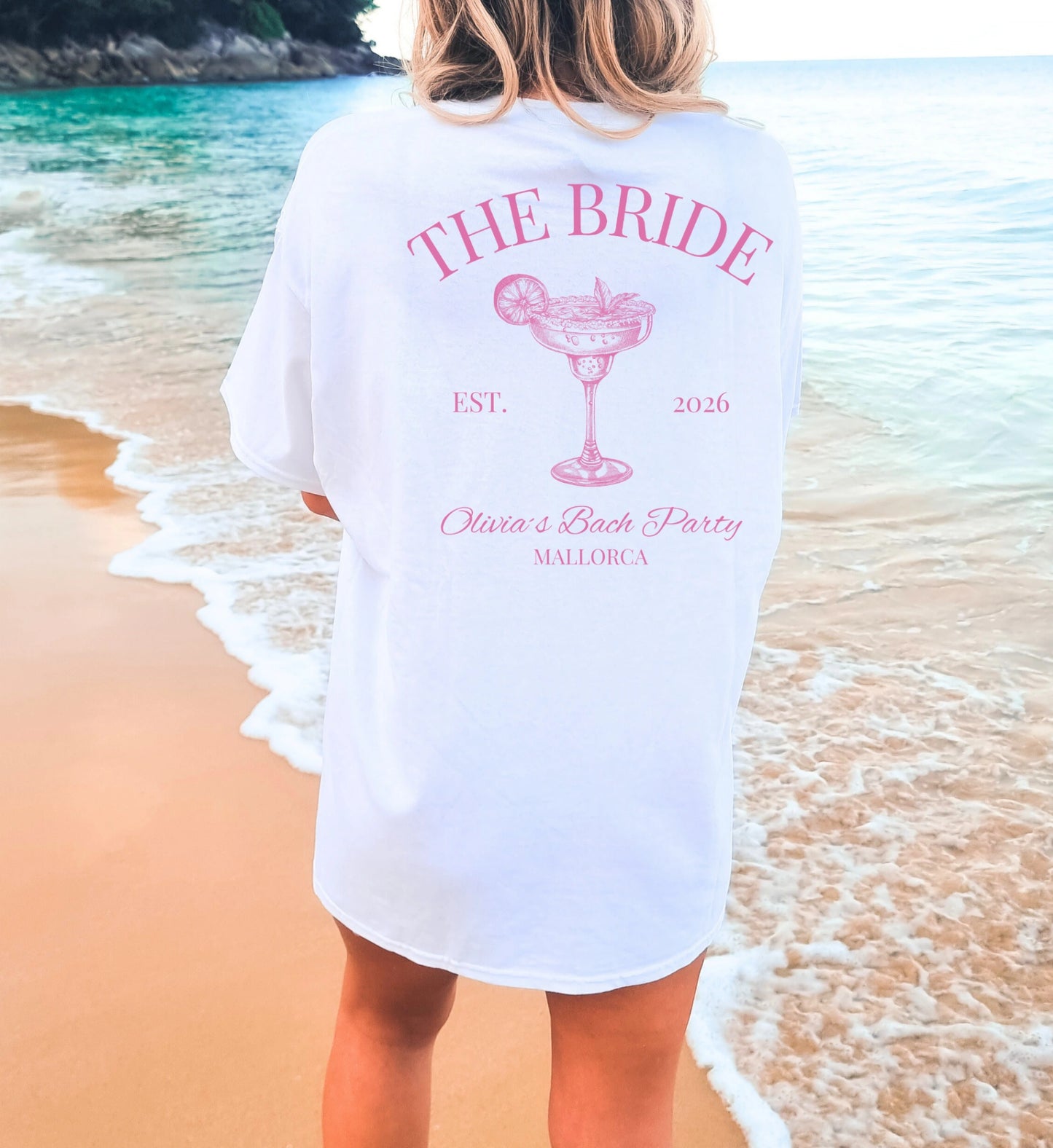 Rückansicht einer Frau am Strand in einem weißen Bachelorette Shirt mit pinkem Margarita-Aufdruck, 'The Bride' Schriftzug und Mallorca-Design für den JGA.