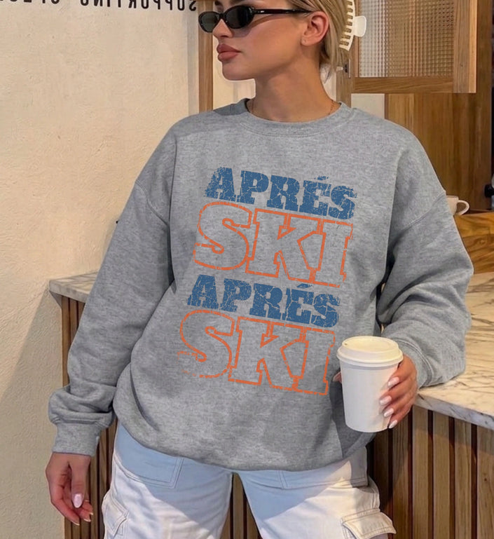 Eine Frau trägt einen grauen, lässigen Apres Ski Sweatshirt mit blau-orangem Vintage-Aufdruck im Used-Look in einer modernen Bar-Umgebung mit Sonnenbrille.