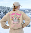 Rückansicht einer Frau in einem beigen Oversized-Sweatshirt mit einem aufgedruckten Motiv einer Maus mit Skibrille, Tutu und Biergläsern vor Bergkulisse.