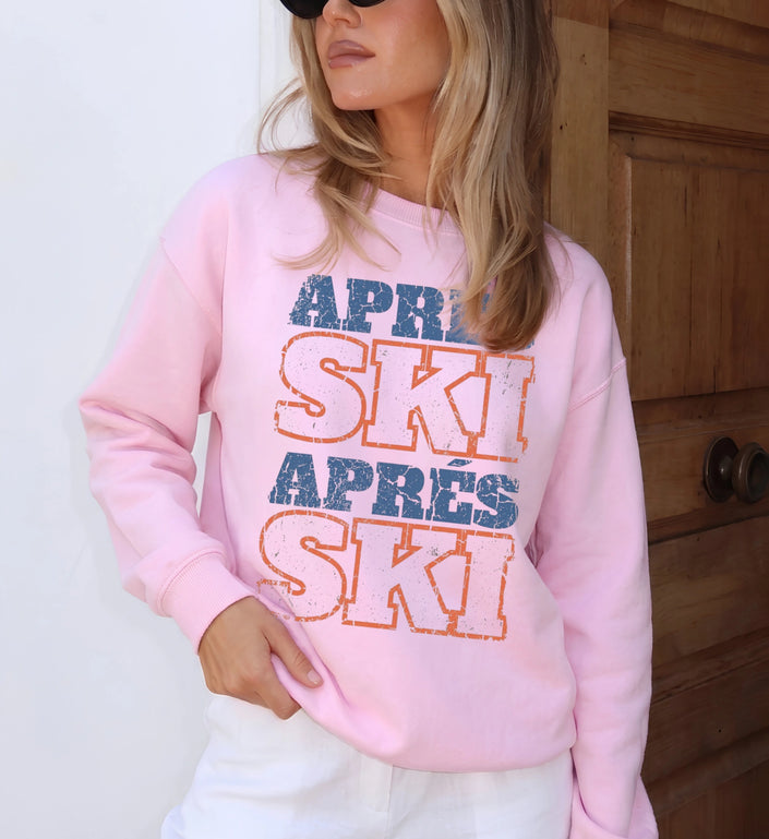 Frau mit blonden Haaren präsentiert einen hellrosa Apres Ski Sweatshirt mit blau-orangem Retro-Frontdruck in einer gemütlichen Innenraum-Kulisse aus Holz.
