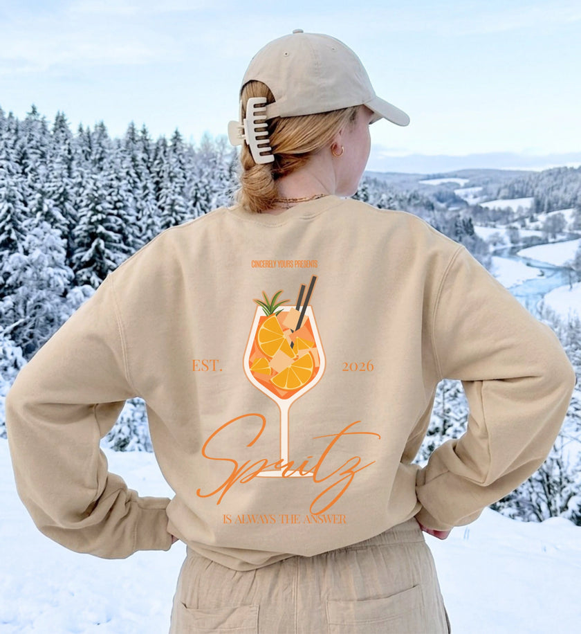 Rückenansicht eines beigen Sweatshirts mit großem Spritz-Cocktail-Motiv und dem Slogan Is always the answer vor einer verschneiten Winterlandschaft.