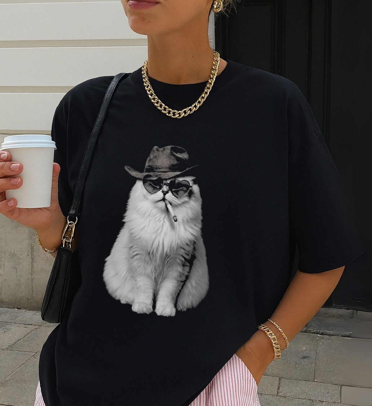 Nahaufnahme eines schwarzen T-Shirts mit Print einer weißen Katze im Cowboy-Look mit Goldkette und Kaffeebecher kombiniert.