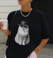 Nahaufnahme eines schwarzen T-Shirts mit Print einer weißen Katze im Cowboy-Look mit Goldkette und Kaffeebecher kombiniert.