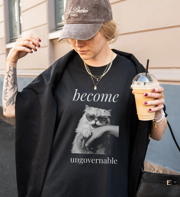 Stylisches schwarzes T-Shirt mit Katzen-Motiv in Herz-Sonnenbrille, getragen von einer Frau mit Baseball-Cap und Eiskaffee in der Hand in einer städtischen Umgebung.
