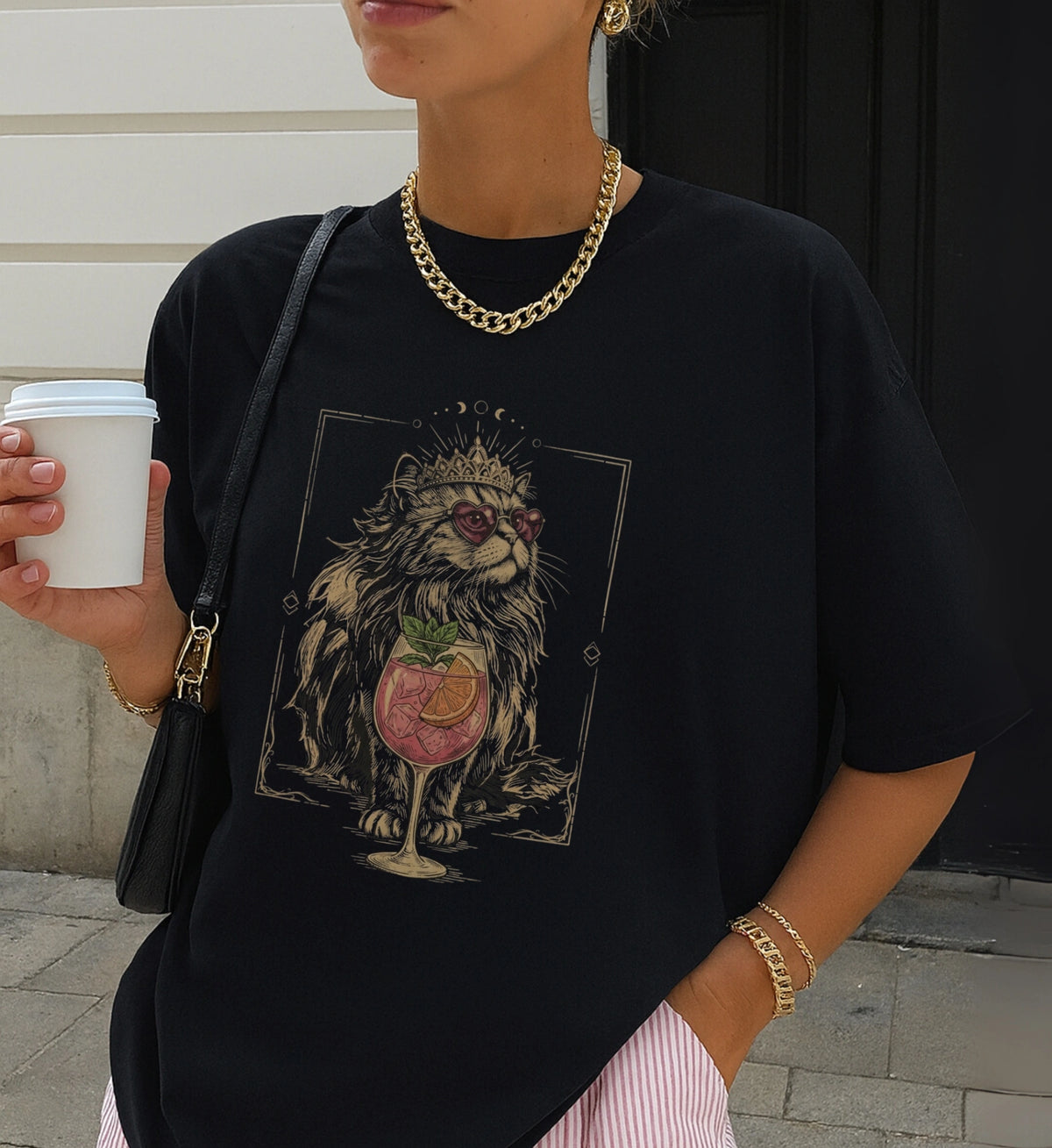 Casual Street-Style Aufnahme einer Frau mit schwarzem T-Shirt, bedruckt mit einer mystischen Katze in einer Krone und einem Spritz-Getränk, kombiniert mit goldener Kette.