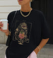 Casual Street-Style Aufnahme einer Frau mit schwarzem T-Shirt, bedruckt mit einer mystischen Katze in einer Krone und einem Spritz-Getränk, kombiniert mit goldener Kette.