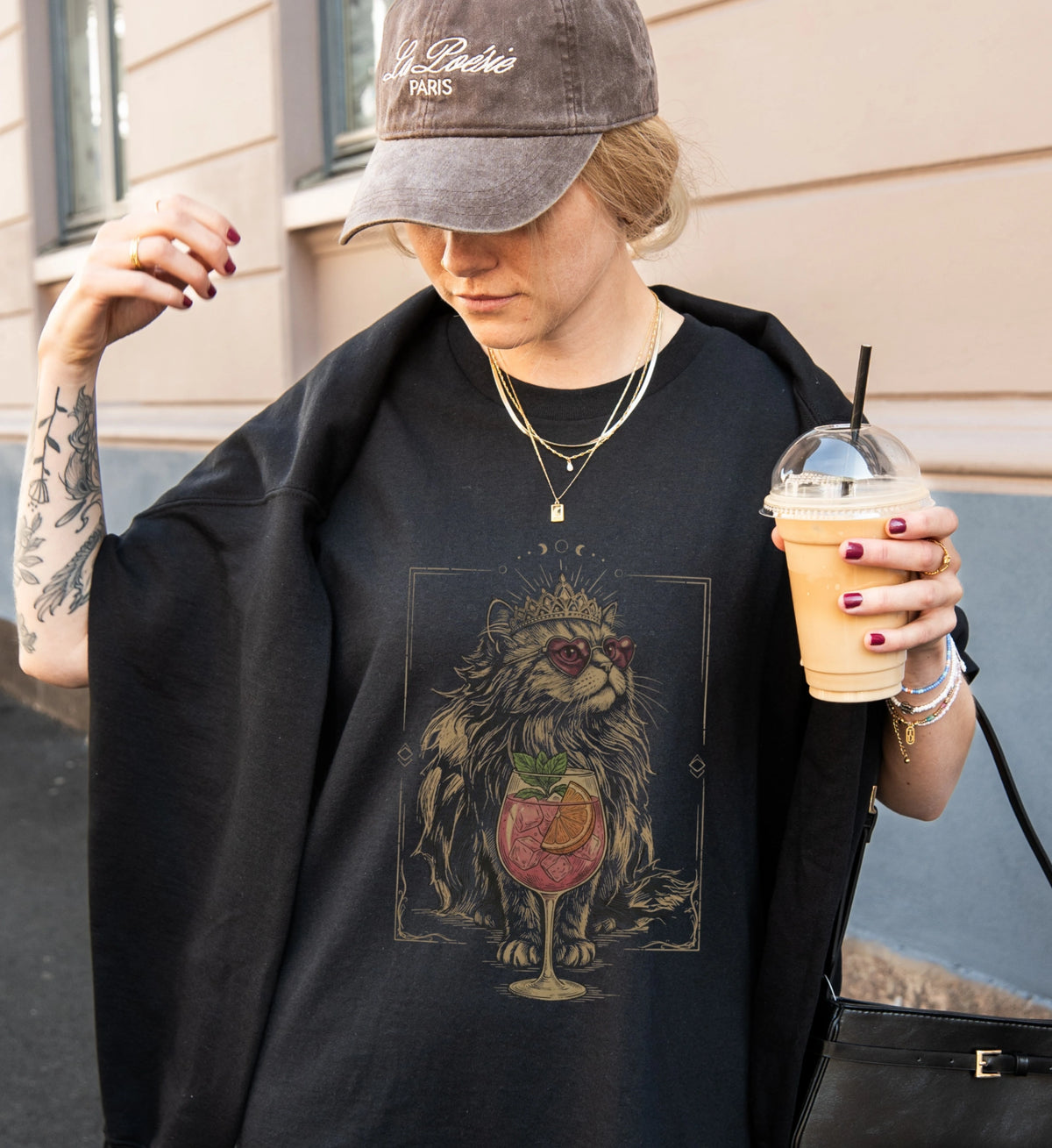 Stylische Frau in einem schwarzen T-Shirt mit mystischem Katzen-Motiv und Cocktail-Grafik, hält einen Iced Coffee in der Hand, trägt eine braune Baseball-Cap und Schmuck.