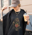 Stylische Frau in einem schwarzen T-Shirt mit mystischem Katzen-Motiv und Cocktail-Grafik, hält einen Iced Coffee in der Hand, trägt eine braune Baseball-Cap und Schmuck.