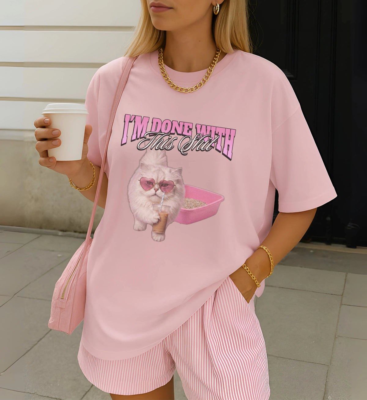 Pastellrosa Cincerely Yours T-Shirt mit unhinged Katzen-Print, Herzbrille und Kaffee, kombiniert mit goldener Kette und gestreiften Shorts im Y2K-Look.