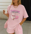 Pastellrosa Cincerely Yours T-Shirt mit unhinged Katzen-Print, Herzbrille und Kaffee, kombiniert mit goldener Kette und gestreiften Shorts im Y2K-Look.
