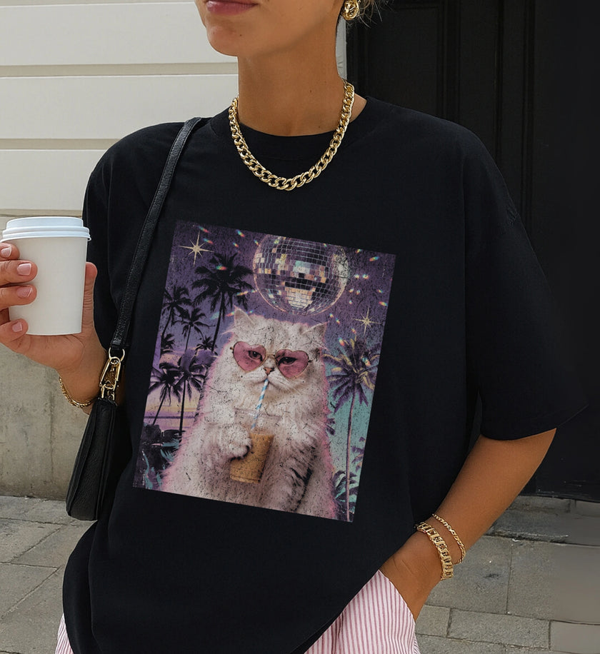 Nahaufnahme einer Frau in schwarzem T-Shirt mit Katze-Grafik, Herz-Sonnenbrille Motiv und Palmen-Hintergrund, hält einen Coffee-to-go Becher in der Hand.