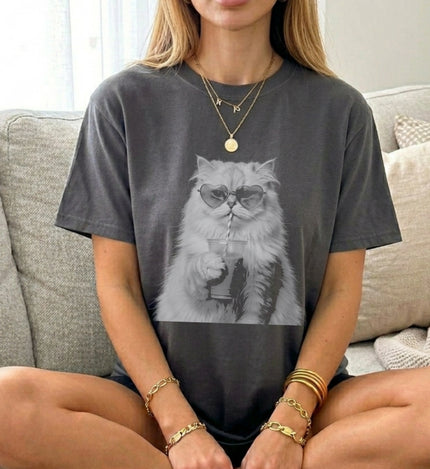 Eine Frau trägt ein anthrazitfarbenes Oversized T-Shirt mit einem Retro-Bootleg-Print einer weißen Katze, die eine Herz-Sonnenbrille trägt und einen Eiskaffee trinkt.