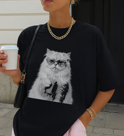 Eine Frau hält einen weißen Coffee-to-go Becher, während sie ein schwarzes T-Shirt mit dem illustrierten Druck einer Katze im Bootleg-Stil trägt.