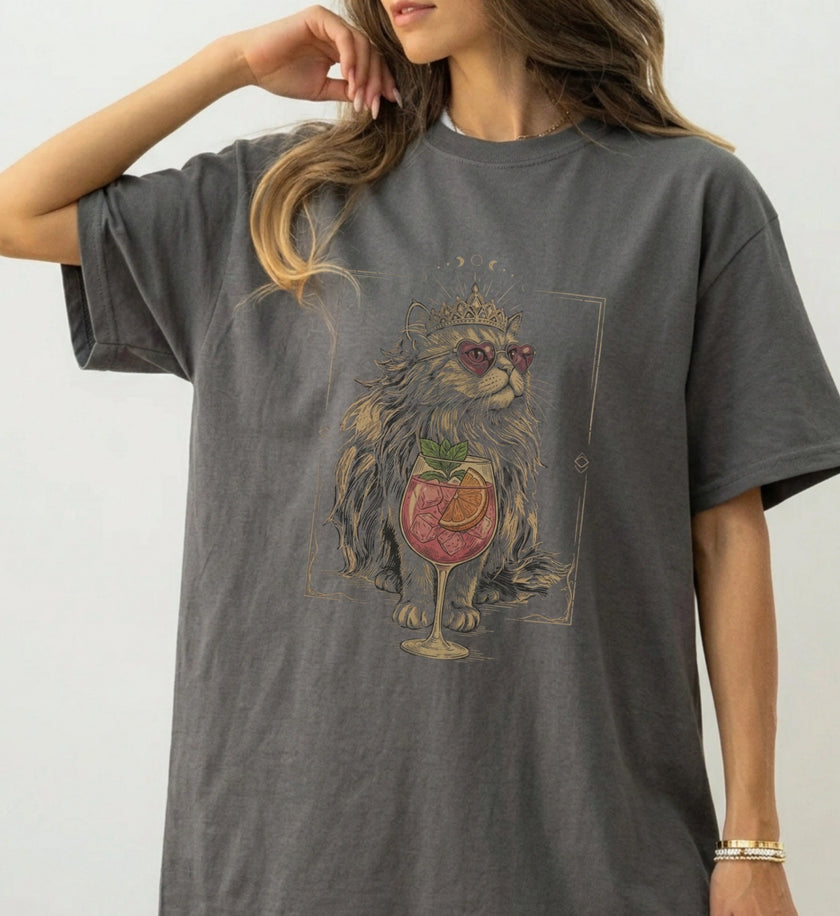 Studioaufnahme einer Frau im grauen Vintage-Look T-Shirt mit detailliertem Print einer edlen Katze mit Krone, Sonnenbrille und einem fruchtigen Cocktail in einem Weinglas.