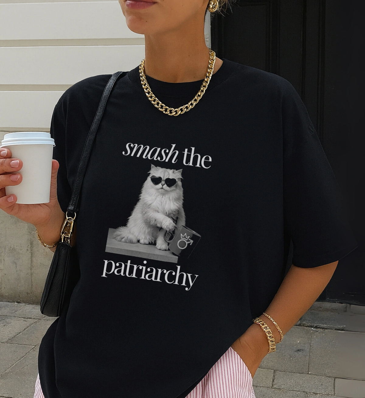 Nahaufnahme eines schwarzen T-Shirts mit Cat-Print und Smash-the-Patriarchy-Text, kombiniert mit goldener Kette und Eiskaffee in einer städtischen Umgebung.