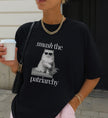 Nahaufnahme eines schwarzen T-Shirts mit Cat-Print und Smash-the-Patriarchy-Text, kombiniert mit goldener Kette und Eiskaffee in einer städtischen Umgebung.
