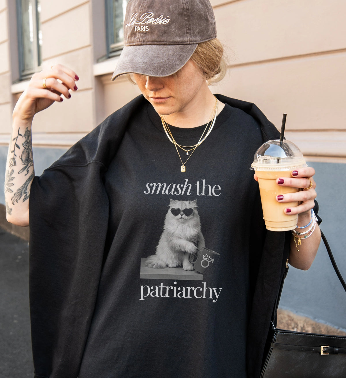 Frau trägt ein schwarzes T-Shirt mit unhinged Cat-Grafik, dazu eine Kappe und einen Eiskaffee, moderner Street-Style Look auf der Straße.