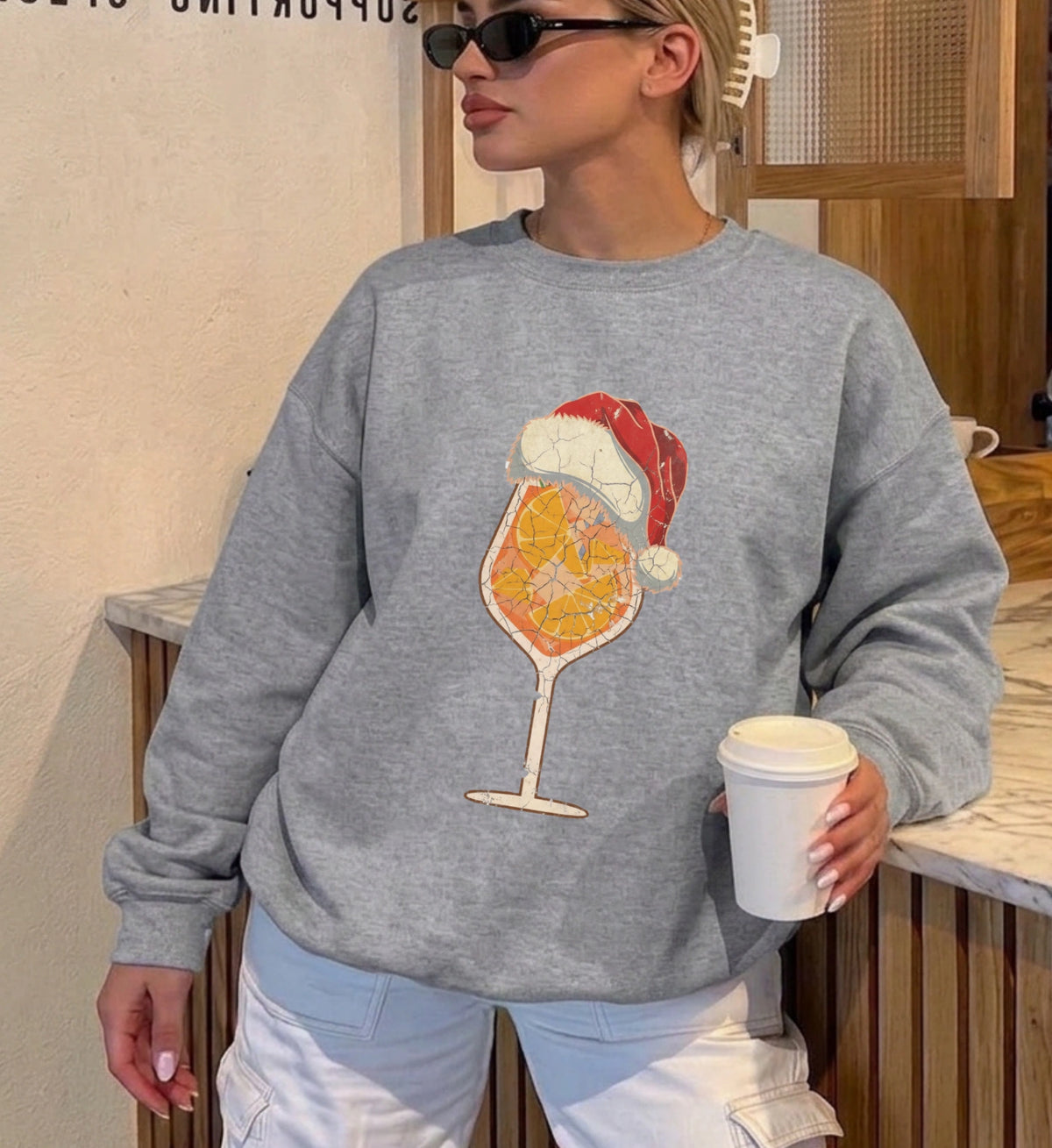 Frau trägt einen grauen Cincerely Yours Christmas Sweater mit aufgedrucktem Spritz-Cocktail Glas, das eine Weihnachtsmütze trägt, Vintage-Grafik-Stil, lässiger Oversize-Schnitt.