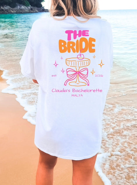 Cincerely Yours Coquette Bachelorette Shirt in Weiß mit neonfarbenem 'The Bride' Aufdruck, Cocktail-Grafik und Schleife, getragen am Strand – ideales JGA Outfit für die Braut.