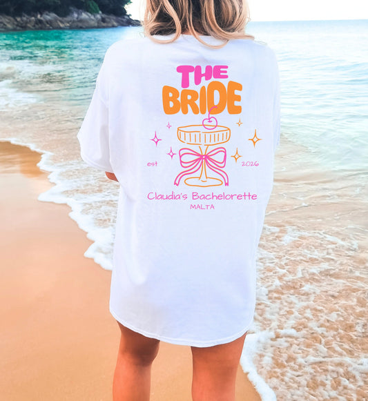 Cincerely Yours Coquette Bachelorette Shirt in Weiß mit neonfarbenem 'The Bride' Aufdruck, Cocktail-Grafik und Schleife, getragen am Strand – ideales JGA Outfit für die Braut.