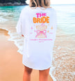 Cincerely Yours Coquette Bachelorette Shirt in Weiß mit neonfarbenem 'The Bride' Aufdruck, Cocktail-Grafik und Schleife, getragen am Strand – ideales JGA Outfit für die Braut.