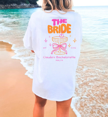 Cincerely Yours Coquette Bachelorette Shirt in Weiß mit neonfarbenem 'The Bride' Aufdruck, Cocktail-Grafik und Schleife, getragen am Strand – ideales JGA Outfit für die Braut.