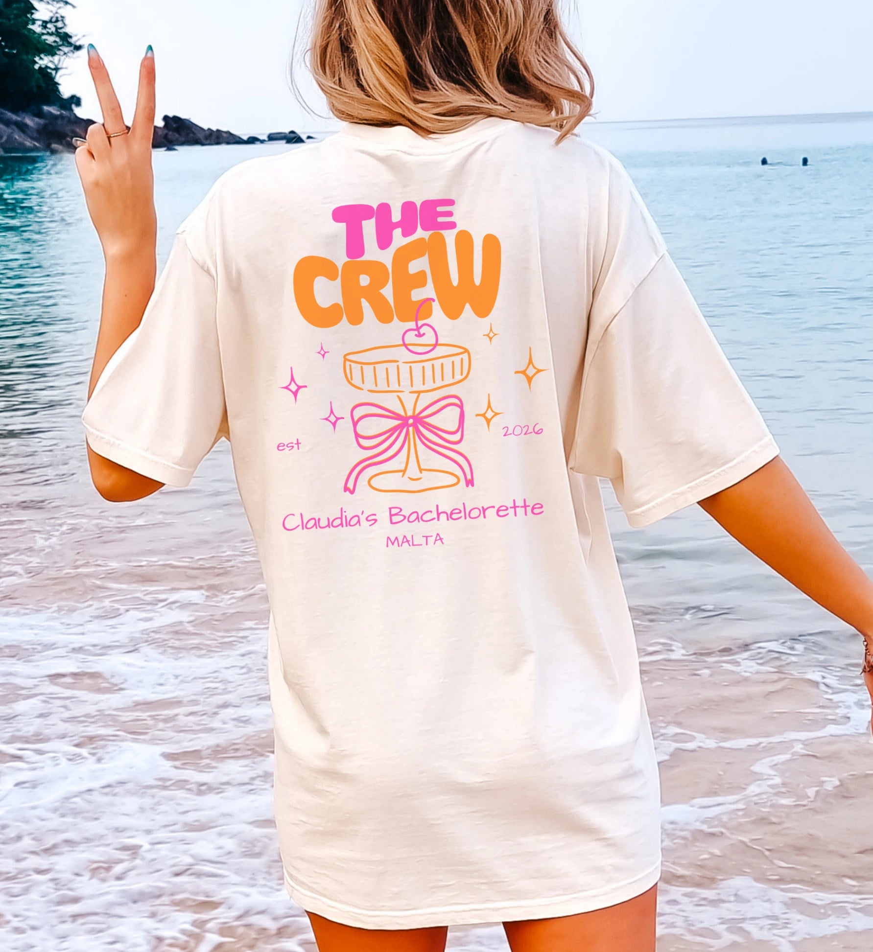 Beiges Cincerely Yours Coquette Bachelorette Shirt mit Neon 'The Crew' Print, Cocktail-Grafik und pinker Schleife für den JGA, getragen am Strand in der Rückansicht.