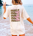 Rückansicht einer Frau am Strand, die ein helles T-Shirt mit großem, mehrzeiligem Malle-Print im Leopard-Design und vertikalem Maus-Schriftzug in Pink trägt.