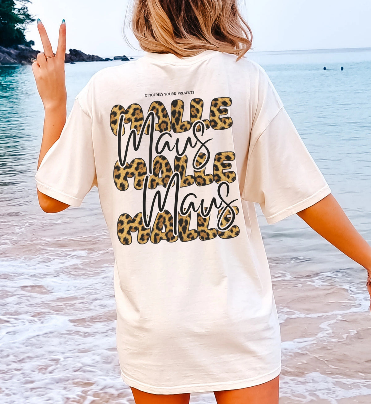 Rückenansicht eines hellen T-Shirts mit großem Mallemaus Backprint in auffälligem Leo-Druck Bubble-Schriftart, getragen von einer Frau am Meeresstrand.
