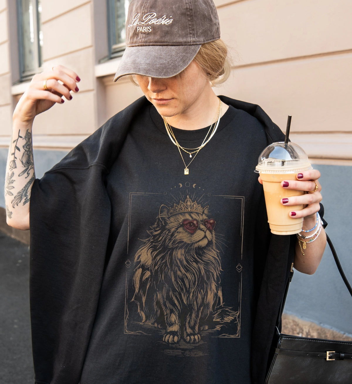 Stylische Frau in schwarzem T-Shirt mit mystischem Katzen-Motiv, trägt eine Cap, hält einen Eiskaffee und präsentiert den modischen Streetwear-Look des Cat-Shirts.