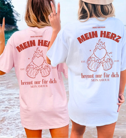 Zwei Frauen am Strand von hinten in rosa und weißen Cincerely Yours Oversized T-Shirts mit dem Rückenprint 'Mein Herz brennt nur für dich' und einer Grafik von anstoßenden Bierkrügen.