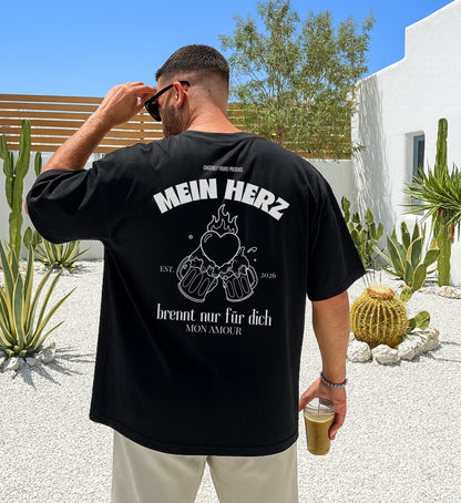 Schwarzes Oversized T-Shirt von Cincerely Yours mit weißem Rückenprint 'Mein Herz brennt nur für dich' und einer Illustration von zwei anstoßenden Bierkrügen mit brennendem Herz, getragen von einem Mann in einer sommerlichen Außenkulisse.