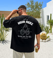 Schwarzes Oversized T-Shirt von Cincerely Yours mit weißem Rückenprint 'Mein Herz brennt nur für dich' und einer Illustration von zwei anstoßenden Bierkrügen mit brennendem Herz, getragen von einem Mann in einer sommerlichen Außenkulisse.