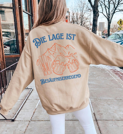Rückenansicht eines beigen Oversized-Sweaters mit blauem Schriftzug und orangefarbener Grafik von Skiern und Bierkrügen im Retro-Stil bei einem winterlichen Straßen-Setting.