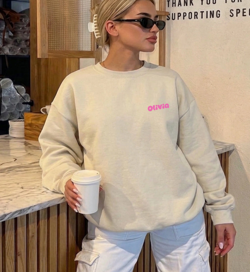 Vorderansicht eines beigen Sweatshirts mit personalisiertem Namen in pinker Bubble-Schrift auf der Brust, getragen von einer Frau in einem Café.
