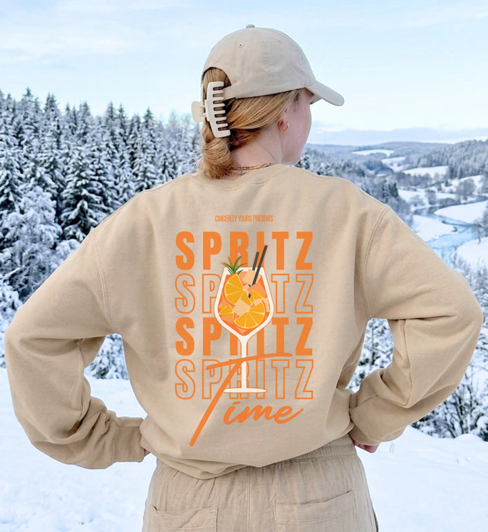 Rückenansicht des beigen Sweatshirts mit großem orangen Spritz Time Cocktail Print vor verschneiter Bergkulisse für Après-Ski und Party.