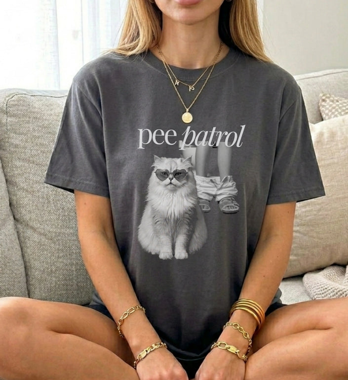Model im dunkelgrauen Pee Patrol Katze T-Shirt im Vintage-Schnitt, sitzend auf einem beigen Sofa mit Goldschmuck.
