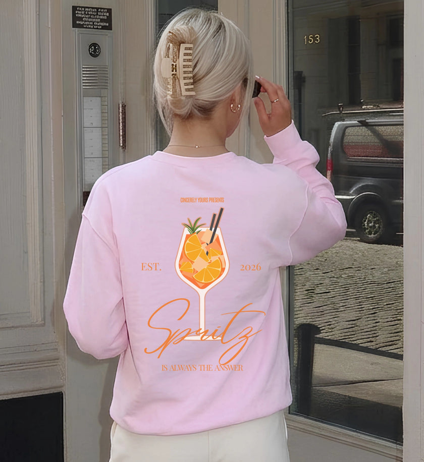 Rückenansicht eines hellrosa Crewneck-Sweatshirts mit aufgedrucktem Sommer-Cocktail-Motiv und dem Text Spritz is always the answer in einer städtischen Umgebung.