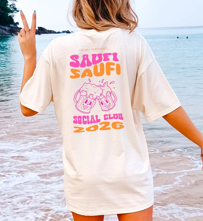 Frau am Strand trägt ein weißes Oversize Party Shirt von Cincerely Yours mit dem Backprint Saufi Saufi Social Club 2026 und zwei lachenden Bierkrügen.